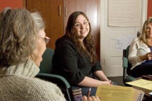 Three women sit in a room having a discussion; one is smiling at the camera while holding papers, and the others are engaged in conversation.