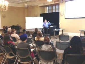 A man stands at the front of a room giving a presentation to a small seated audience, with a whiteboard and projection screen behind him.