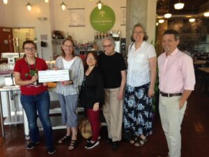 Six people stand together inside a cafe, smiling at the camera; one person holds an envelope and flowers. A sign reading "Creamery Cafe" is visible in the background.