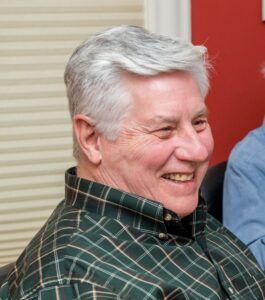 An older man with short gray hair and a plaid shirt is sitting indoors, smiling and looking to the right.