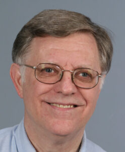 A middle-aged man with gray hair and glasses smiling at the camera, wearing a light blue collared shirt, against a plain gray background.