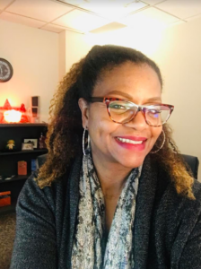A woman with glasses and curly hair smiles at the camera in an office with shelves and a salt lamp in the background.