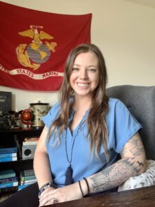 A woman with long hair and tattoos sits smiling at a desk; a red United States Marine Corps flag hangs on the wall behind her.