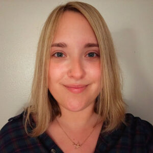 A woman with straight blonde hair and a nose stud, wearing a dark plaid shirt and a gold necklace, smiles at the camera against a plain light background.