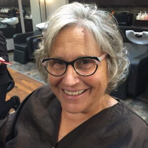 Smiling older woman with short gray hair and glasses sits in a salon chair, wearing a brown cape, with salon sinks and chairs visible in the background.