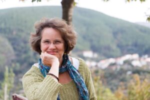 A woman with glasses and a blue scarf sits outdoors, resting her chin on her hand, with forested hills and a town visible in the background.