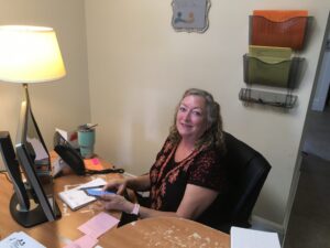 A woman sits at a desk using her phone, with papers, a lamp, and office supplies around her. A wall organizer with colored folders is mounted nearby.
