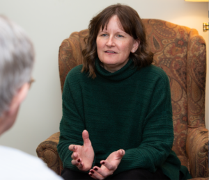 A woman in a green sweater sits in an armchair, gesturing with her hands while talking to another person whose back is visible in the foreground.