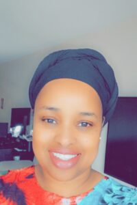 A person wearing a black headwrap and a colorful top smiles at the camera indoors, with a TV and furniture visible in the background.