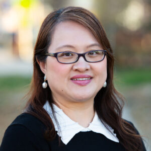 A woman with long brown hair and glasses, wearing a black top with a white collar, smiles outdoors with a blurred background.