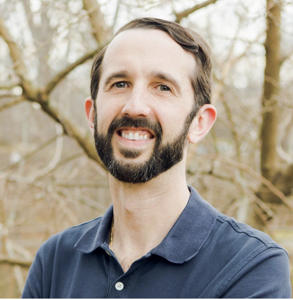 A man with a beard and short brown hair, wearing a navy blue collared shirt, stands outside in front of leafless trees, smiling at the camera.
