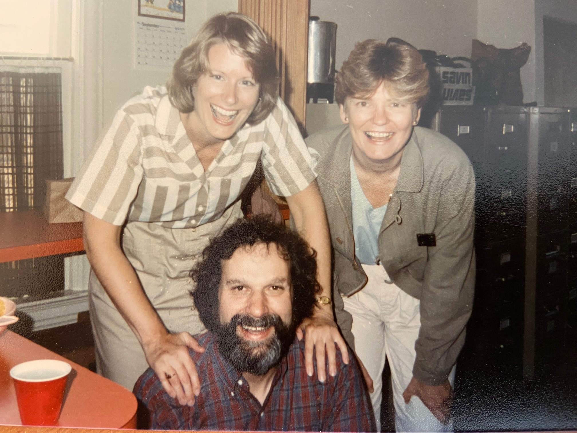 Three people pose and smile indoors; two women stand behind a seated bearded man. There are filing cabinets and a calendar in the background.
