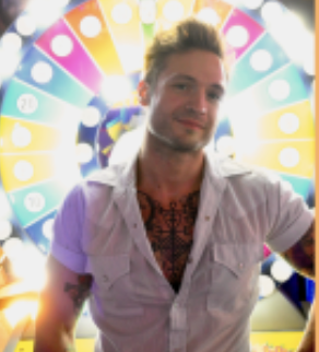 A man with tattoos and a white shirt stands in front of a colorful, illuminated prize wheel at an arcade or carnival.