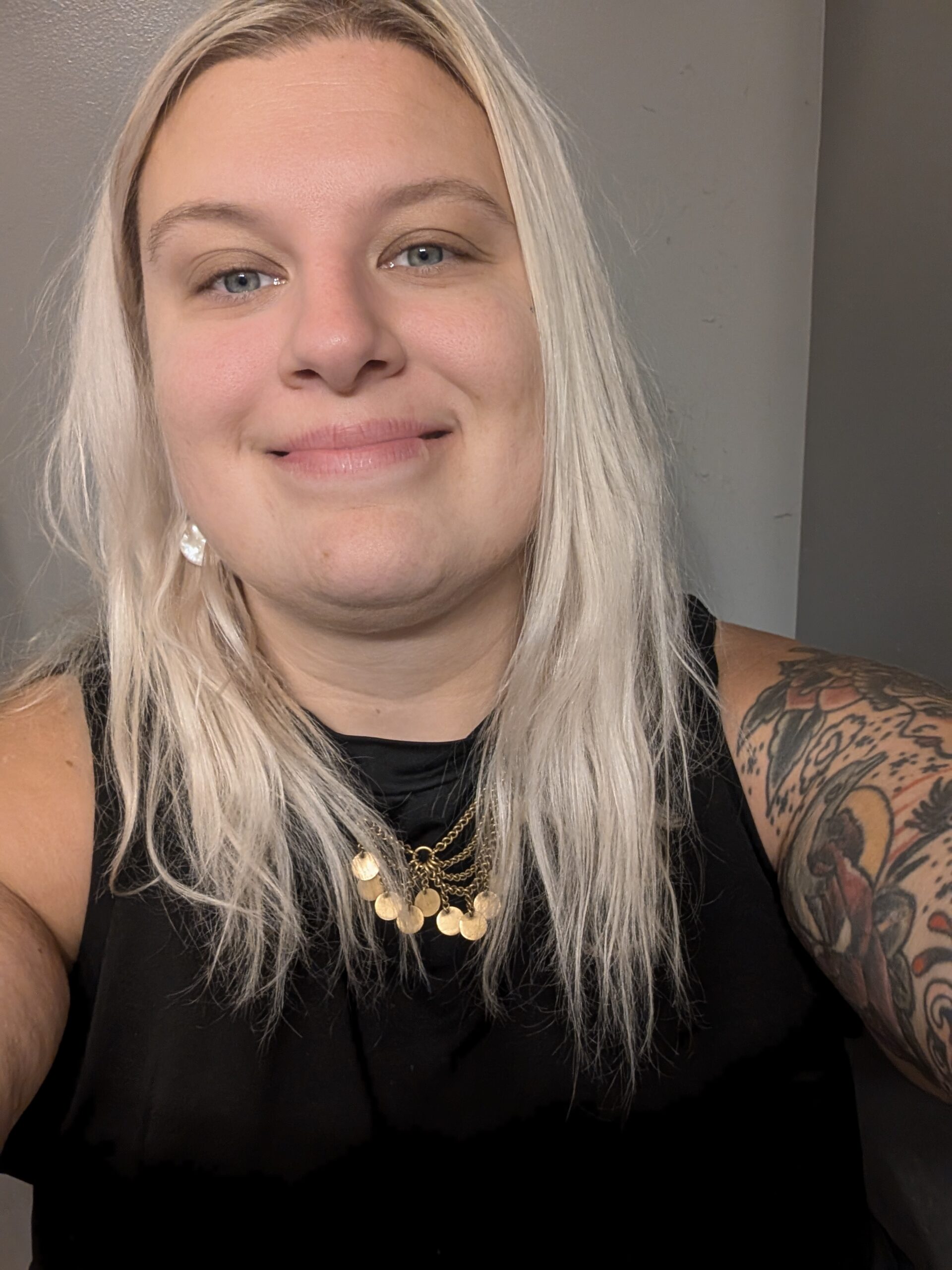 A person with long, light blonde hair and a floral tattoo on their arm smiles at the camera, wearing a black sleeveless top and layered gold necklaces.