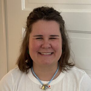 A woman with shoulder-length brown hair and a white shirt smiles at the camera, wearing a blue necklace with three decorative beads. She is in front of a white door.
