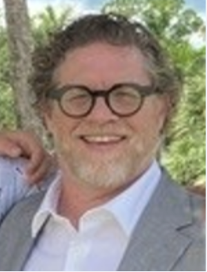 A man with curly hair, round glasses, and a beard is wearing a light grey suit and white shirt, smiling outdoors with greenery in the background.