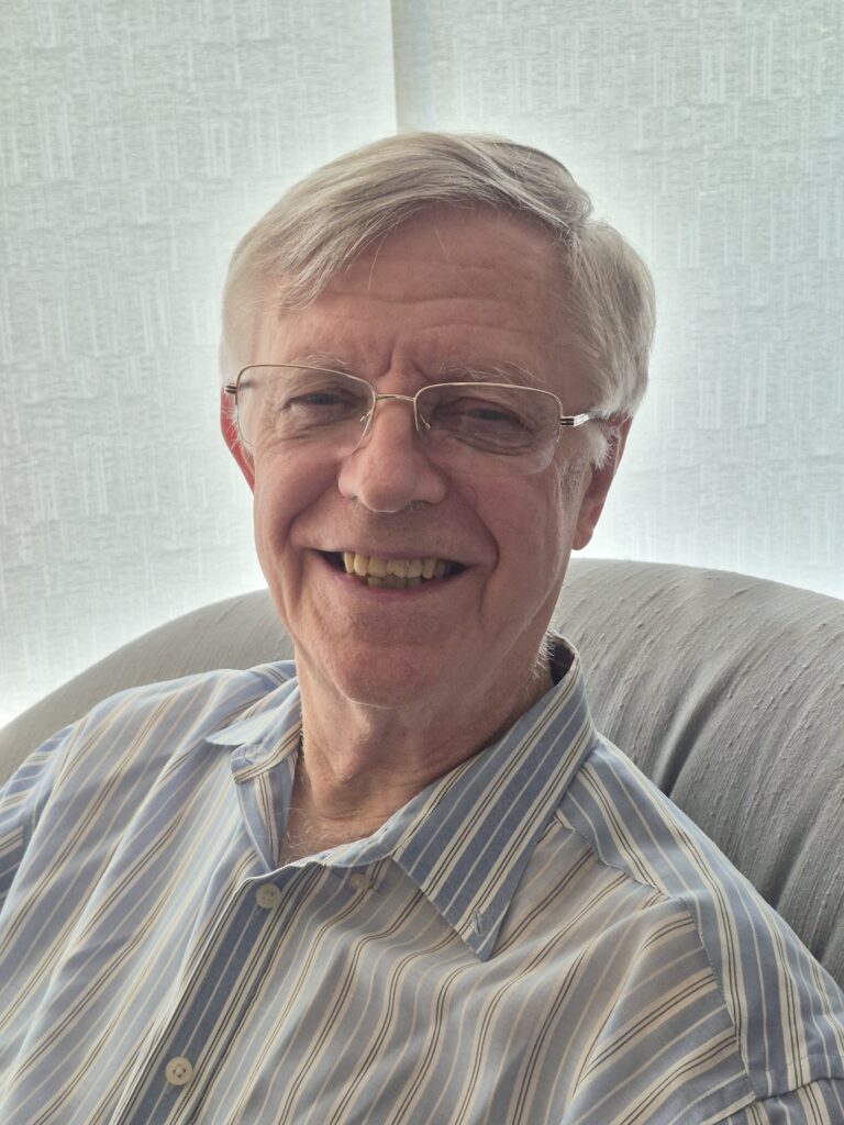 Older man with glasses and gray hair, smiling, wearing a blue and white striped shirt, seated in a softly lit room with light-colored curtains in the background.
