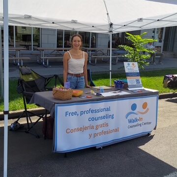 A woman stands behind a table under a canopy offering free, professional counseling at the Walk-In Counseling Center, with bilingual signs in English and Spanish.