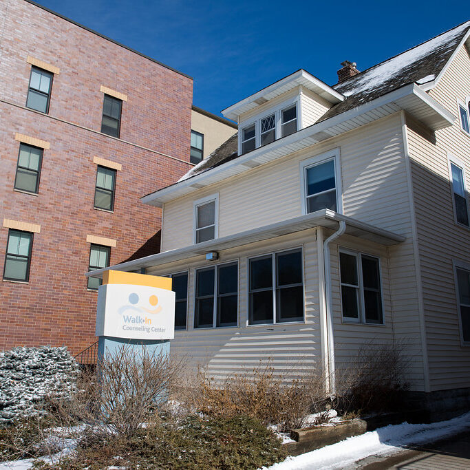 A beige two-story house with a sign reading “Walk-In Counseling Center,” next to a tall brick building on a sunny winter day.
